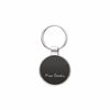 A round black metal keychain with a silver-tone border and key ring, featuring the white script engraving 'Pierre Cardin' on the front, set against a plain white background.