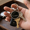 A close-up of a hand holding a keychain with a round black tag engraved with 'Pierre Cardin' in silver-tone lettering, attached to metal key rings and several keys.
